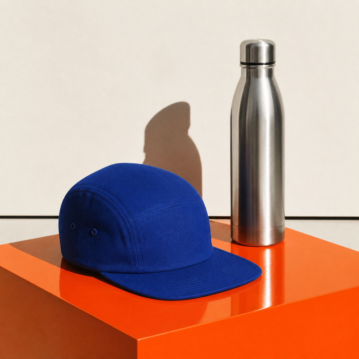 A blue five panel cap and silver bottle on an orange plinth.
