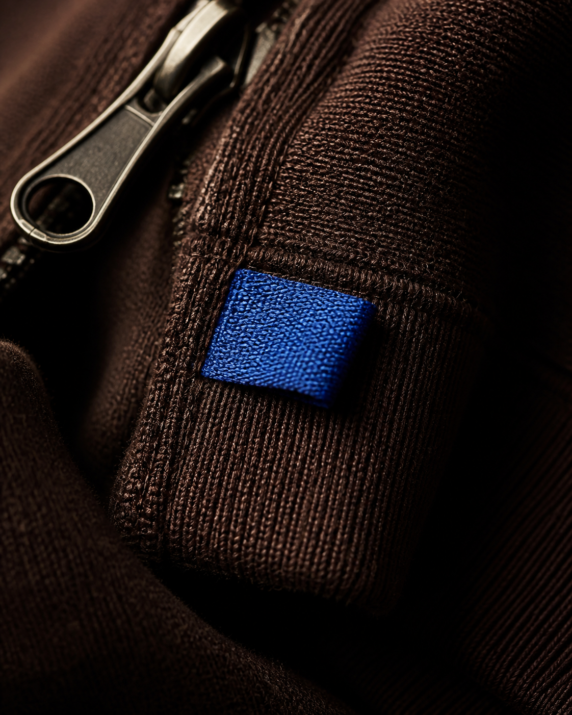 Close detail of dense brown garment fabric, stitchwork and a cobalt label.